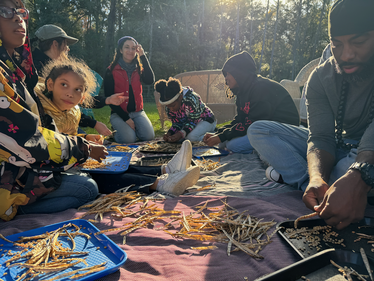 A group of young students and adults sit on the ground processing seeds.